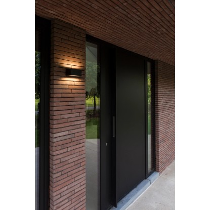 Modern LED kültéri fali lámpa egy sötét fém bejárati ajtó mellett egy téglafalon. Modern LED kültéri fali lámpa egy sötét fém bejárati ajtó mellett egy téglafalon.