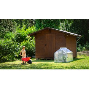 Vitavia Merlin mini üvegház a kerti ház mellett, gyerek talicskával a kertben.