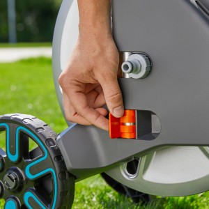 Közelkép: Kéz működteti a narancssárga mechanizmust a Gardena Aqua Roll M tömlőkocsi készleten.