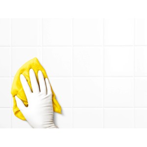 Kéz fehér, latexmentes eldobható kesztyűben fehér csempét tisztít sárga ruhával.